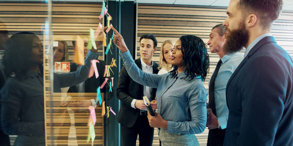 A diverse group of individuals gathered around a glass wall covered in colorful sticky notes.