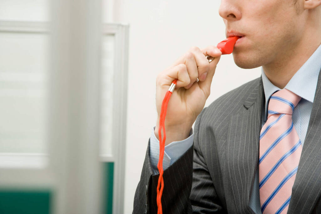 man in a grey suit with a pink and blue striped tie holds a plastic orange whistle in his mouth